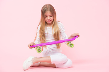 A skater girl or skateboarder. Little girl holding skateboard on pink background. Cute small girl with violet penny board. Adorable child with skater girl style and look