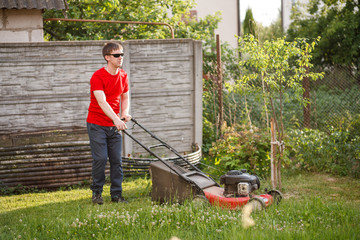 Young man in red t-shirt and sunglasses mows the grass with a grass-cutter on the lawn in the...