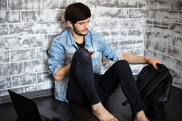 young brunette man sitting on the floor with laptop and smartphone on the background of grey brick wall 