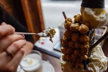 Woman eating Dessert based on vanilla ice cream and chocolate syrup with cookies, marshmallows, popcorn, cream in glass with at wooden table in cozy cafe. Crazy freakshare food trend.