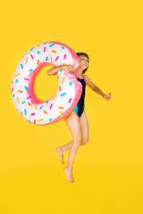 Obraz na płótnie Canvas Cute girl in blue swimsuit with huge nflated rubber ring jumping on yellow colour background