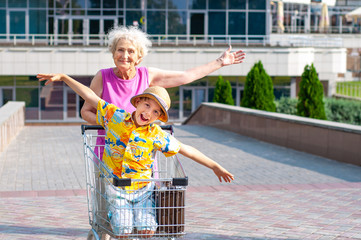 A cheerful grandmother rolls her happy grandson in a basket trolley near shops in the city...