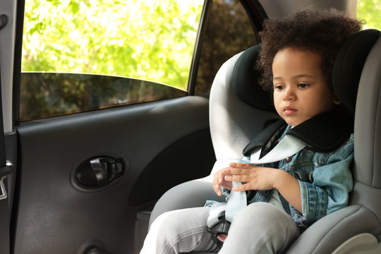 Cute African-American girl sitting in safety seat alone inside car. Child in danger
