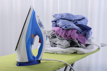 Modern electric iron and heap of clothes on board indoors. Laundry day
