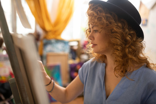 Curly red-haired artist wearing black hat standing near canvas