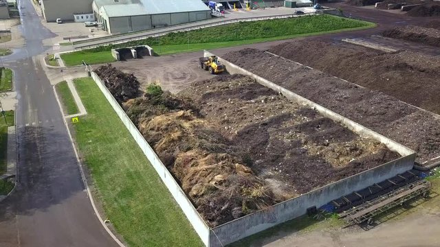 Drone shot of industrial dunghills near some buildings, surrounded by green fields, with some birds flying around, on a sunny day