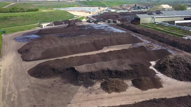 Drone shot of industrial dunghills near some buildings, surrounded by green fields, with some birds flying around, on a sunny day