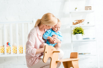 blonde mother holding in arms cute toddler son near rocking horse