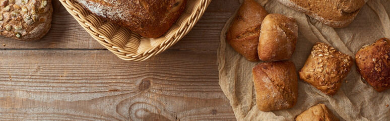 top view of fresh baked bread in wicker basket and buns on cloth on wooden table, panoramic shot