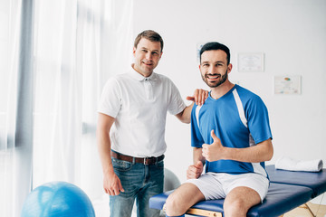 smiling Physiotherapist near happy football player showing thumb up in hospital