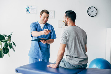 smiling Physiotherapist with diagnosis and pen gesturing near patient in hospital