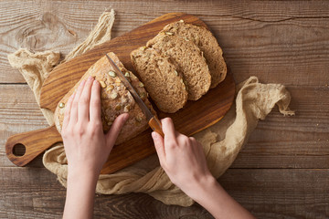 top view of woman cutting bread with seeds on wooden chopping board