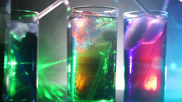 Multicolored glasses filled with alcoholic drinks, with splases of ice cubes falling inside, standing on the mirror surface. Black background. Conceptual, celebrated, commercial design. Closeup.