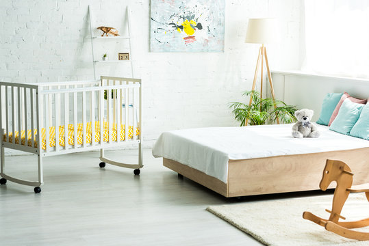 baby crib near bed with white bedding and pillows near teddy bear and wooden rocking horse