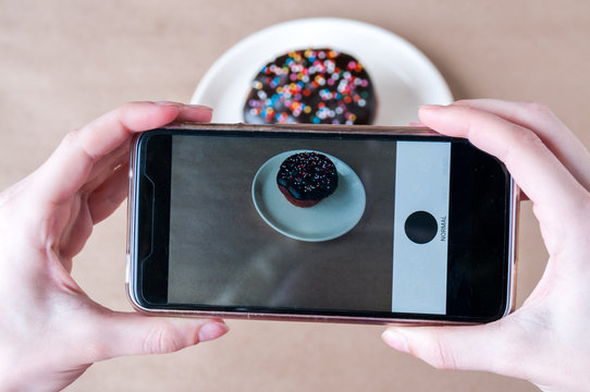 Women's hands over the table holding a smartphone and take pictures of a chocolate donut on a plate