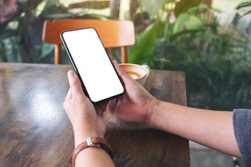Mockup image of hands holding black mobile phone with blank desktop screen with laptop and coffee cup on the table