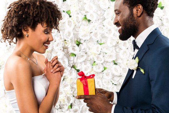 happy african american man giving present to surprised bride near flowers