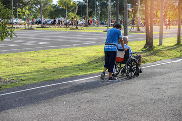 The old man on a wheelchair and his adult son are walking in the park. A man is assisting his father.