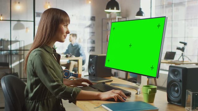 Beautiful Brunette Female Video Editor Works on Her Personal Computer with Big Green Screen Mock Up Display. She Works in a Cool Office Loft. Other Male Creative Colleague Walks in the Background.
