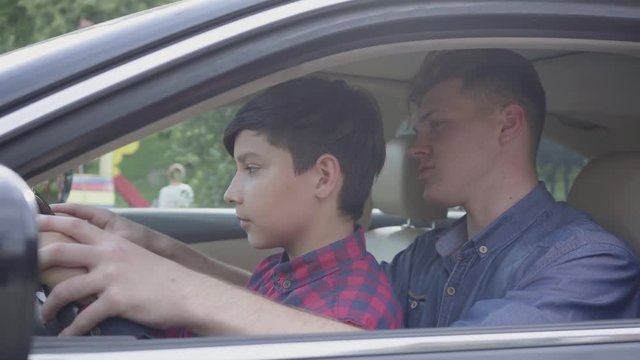 Young Father Teaching His Son To Drive The Car Close Up. The Boy Sitting On Father's Laps, Listening To Him. The Child And Father Spending Time Together. A Man Passes On Knowledge To Son