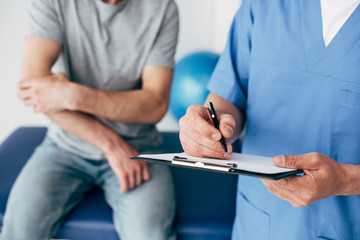 Cropped view of doctor writing on clipboard and patient touching arm in massage cabinet at clinic