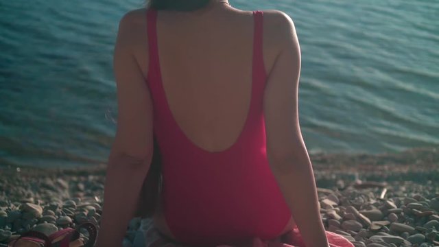 A beautiful young girl in a red one-piece swimsuit sits on the seashore in the sunset sun. The girl sits back in the frame.