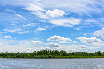 view from the river to the wooded shore. Blue sky with clouds on summer day