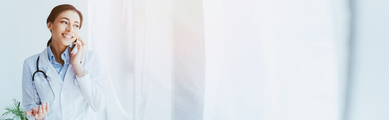 panoramic shot of smiling latin doctor talking by smartphone and looking away
