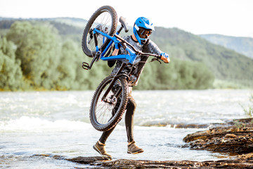 Professional well-equipped cyclist carrying bicycle on the rocky riverside in the mountains. Concept of a freeride cycling