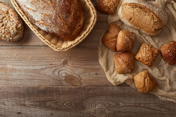 top view of fresh baked bread in wicker basket and buns on cloth on wooden table