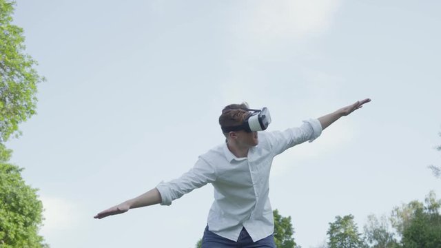 Young man in virtual reality headset spread his arms to the side, imitating the flight of an airplane in the park enjoying the realistic image. Modern technologies. The guy playing a video game