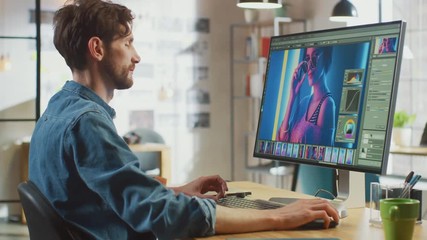 Male Digital Editor in Jeans Shirt Works in Photo Editing Software on His Personal Computer with Big Display. He Works in Cool Office Loft. Other Female Creative Colleague Walks in the Background.