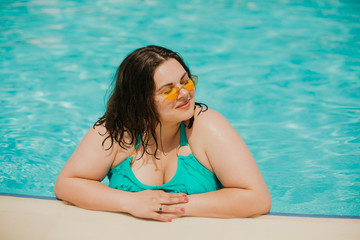 Attractive busty curvy woman in yellow sunglasses and a blue swimsuit resting by the pool. Stylish accessories, fringe, fashion for plus size. Bodypositive, natural real beauty, resort, summer