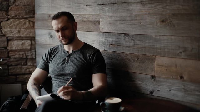 Brutal man with bristles and tattooed hands sits at a table in a cafe and takes notes in his notebook on the background of a beautiful wooden plank wall. Left-handed guy makes notes on paper