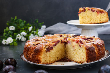 Plums pie with a cut out piece is located on a plate on a dark background, piece of pie in the background