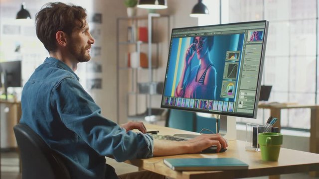 Male Digital Editor in Jeans Shirt Works in Image Editing Software on His Personal Computer with Big Display. He Works in Cool Office Loft. Other Female Creative Colleague Walks in the Background.