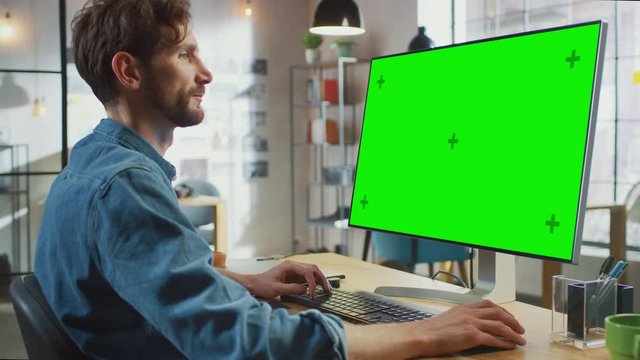 Male Creative Designer with Beard and Jeans Shirt Works on His Personal Computer with Big Green Screen Mock Up Display. He Works in a Cool Office Loft. Female Creative Colleague Walks in Background.