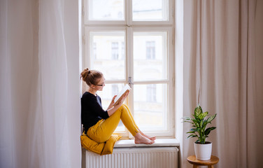 A young female student with a book sitting on window sill, studying. - obrazy, fototapety, plakaty