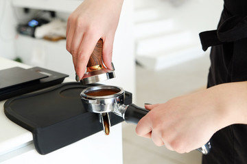 Close up shot of female barista wearing black stylish apron using portafilter coffee machine equipment to press grounded beans near coffee machine in cafe place. Close up, copy space, background.