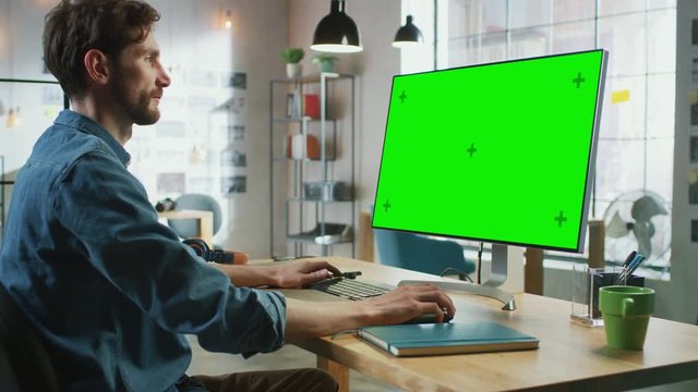 Young Professional Creative Employee Sits Down and Works on His Personal Computer with Big Green Screen Mock Up Display. He Works in a Cool Office Loft. Other Female Colleague Walks in the Background.