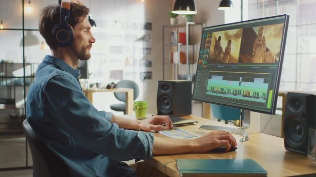 Male Video Editor Puts Headphones and Starts Working with Footage on His Personal Computer with Big Display. He Works in a Cool Office Loft. Creative Man Wears a Jeans Shirt.