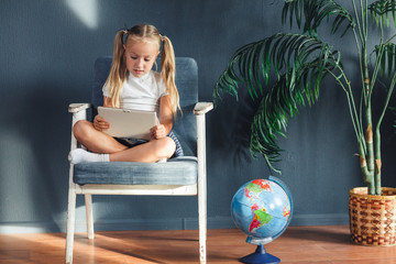 Pretty smiling blondy girl relaxing on a chair near the globe indoors at home with a tablet pc in her socks and jeans.