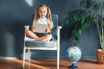 Pretty smiling blondy girl relaxing on a chair near the globe indoors at home with a tablet pc in her socks and jeans.