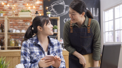 Attractive young waitress talking with regular customer friend. girl client using mobile phone chatting with barista while take order in coffee shop. two smiling women friendship having conversation.