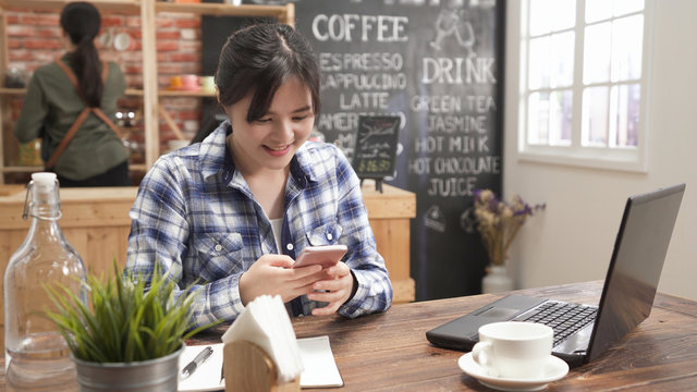 asian business woman in casual wear typing text message on smart phone in cafe bar. young girl freelancer sitting at wooden table with coffee using mobile phone in shop. back view waitress in counter
