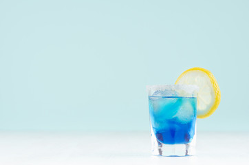 Tropical fresh alcohol cocktail with blue curacao liqueur, ice cube, sugar rim, lemon in misted shot glass on soft light white, pastel green background.