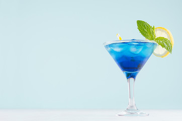 Exotic cold shot glass cocktail with blue curacao, ice cubes, lemon slice, yellow straw, green mint on soft light mint color background and white wood board.