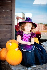 Happy Halloween! Cute cheerful little witch with a magic wand. Beautiful child girl in witch costume sitting on the big pumpkin, conjuring and laughing.