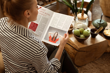 Curious young blonde lady in striped jacket inspecting female magazine