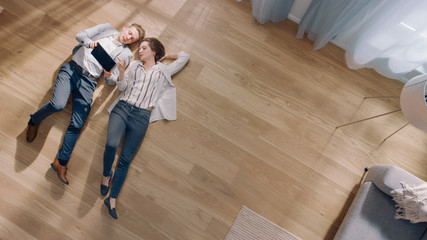 Young Couple are Lying on a Wooden Flooring in an Apartment. They are Using a Tablet Computer. Cozy Living Room with Modern Interior, Grey Sofa and Wooden Parquet. Top View Camera Shot.